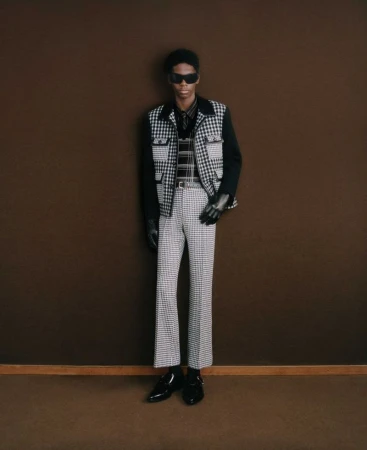 Fashionable African-American male model poses in black and white houndstooth vest and pants against a brown wall in a studio setting.