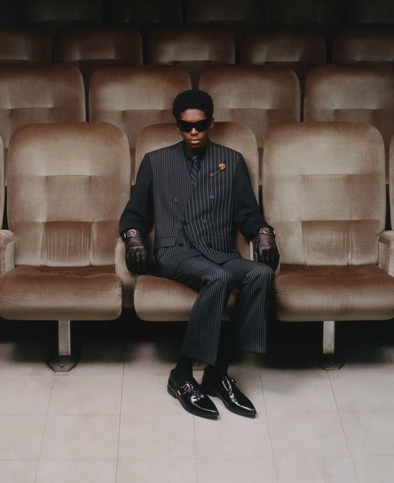Man in a pinstripe suit seated in a vintage theater chair, wearing sunglasses and gloves, striking a poised, elegant pose.