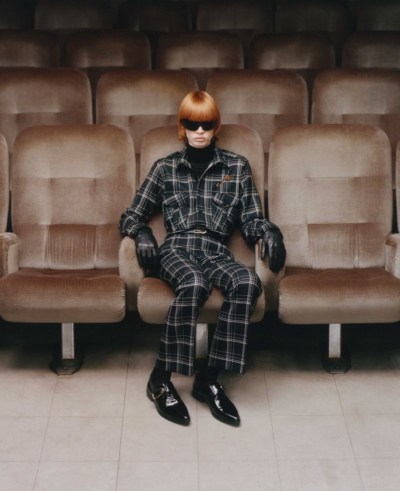 A person with orange hair sits in an empty movie theater, wearing sunglasses, a plaid outfit, and black leather shoes, gazing directly at the camera.