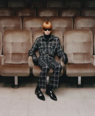 A person with orange hair sits in an empty movie theater, wearing sunglasses, a plaid outfit, and black leather shoes, gazing directly at the camera.