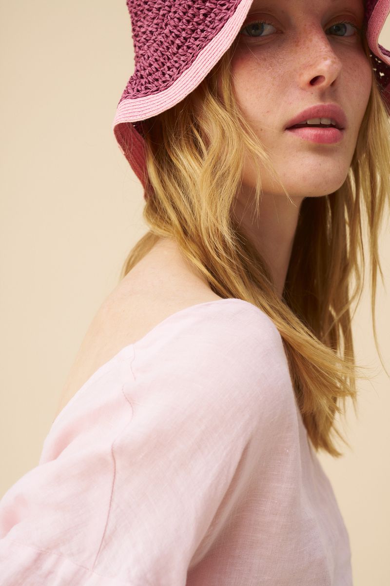 Woman wearing a pink and purple raffia hat and a pink linen top, with fair skin and blonde hair, posed against a neutral backdrop.