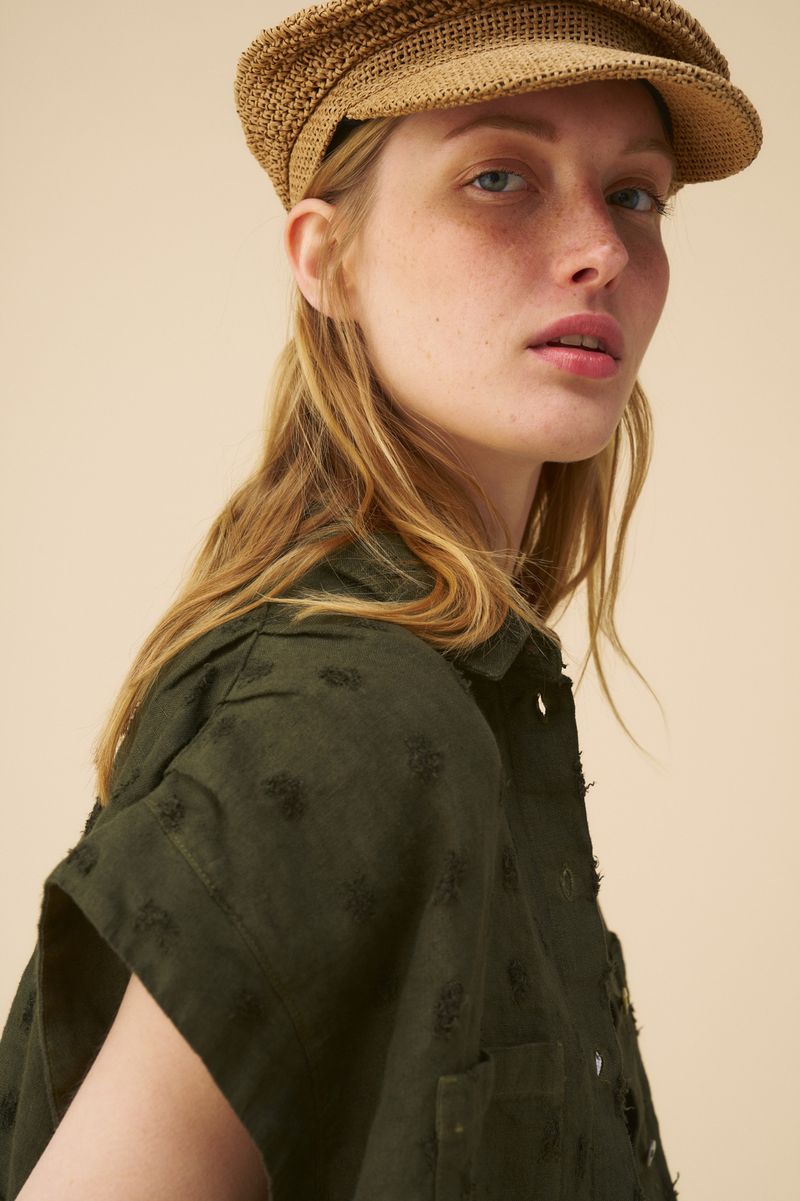 Close-up of a blonde woman wearing a woven cap and green shirt with short sleeves in front of a beige backdrop. Fashion portrait.