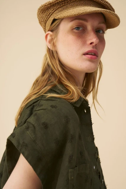 Close-up of a blonde woman wearing a woven cap and green shirt with short sleeves in front of a beige backdrop. Fashion portrait.