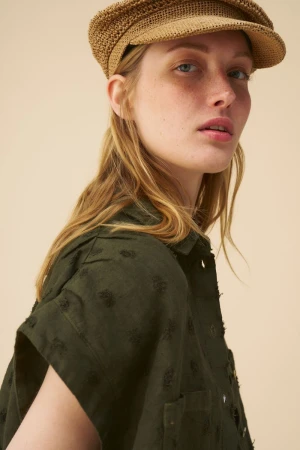 Close-up of a blonde woman wearing a woven cap and green shirt with short sleeves in front of a beige backdrop. Fashion portrait.