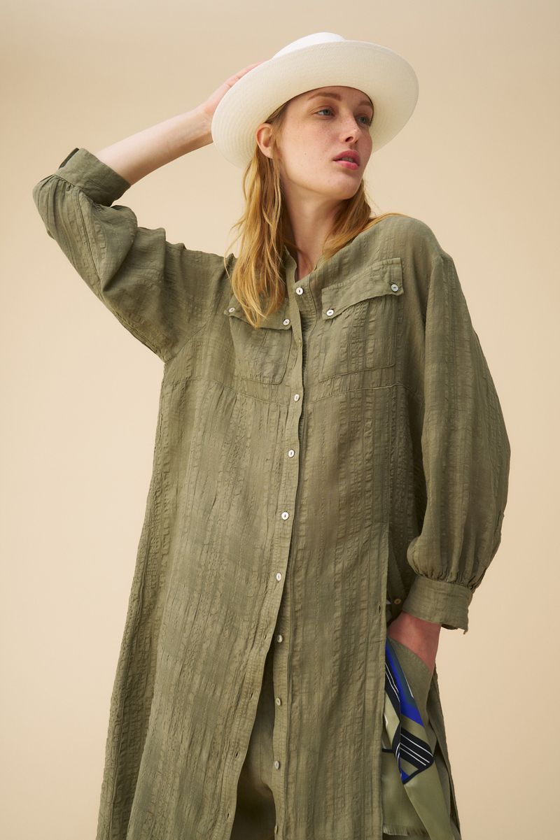 A fair-skinned adult female poses in a button-down olive-green dress and a white hat in a studio setting with neutral background.