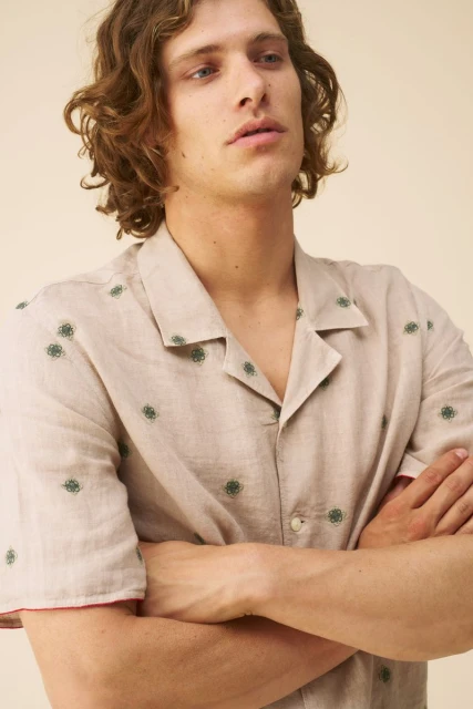 Portrait of a young adult male with wavy hair, wearing a patterned shirt with short sleeves, posing with arms crossed in a studio setting.