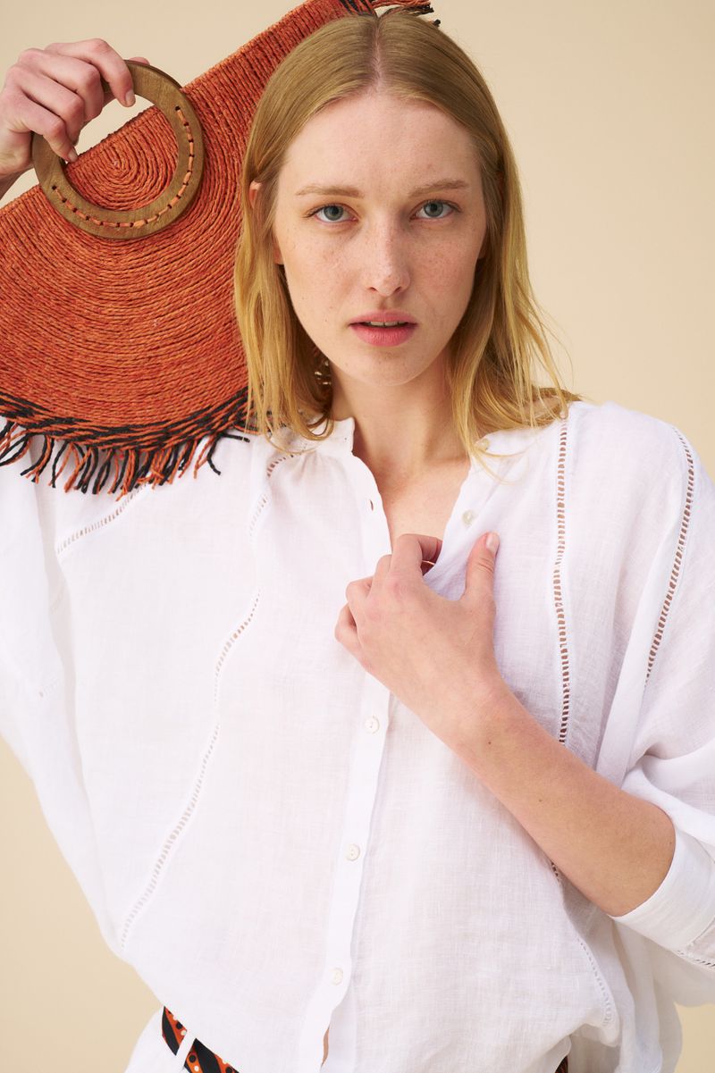 Woman poses with a straw tote bag in a studio, wearing a white linen shirt. Close-up studio shot with neutral background.