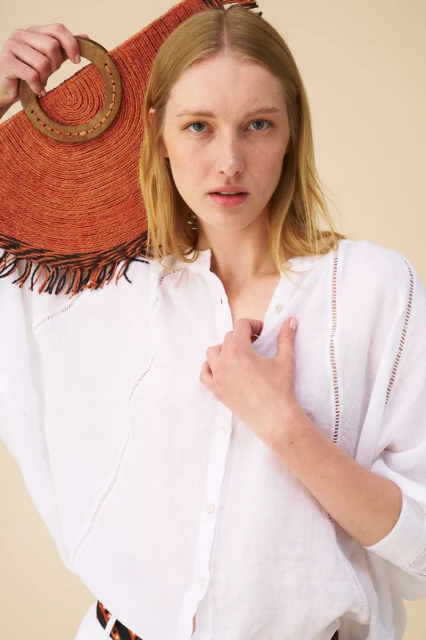 Woman poses with a straw tote bag in a studio, wearing a white linen shirt. Close-up studio shot with neutral background.
