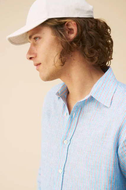 Profile shot of a light-skinned adult male in a blue-striped shirt and white baseball cap, indoors against a neutral backdrop.