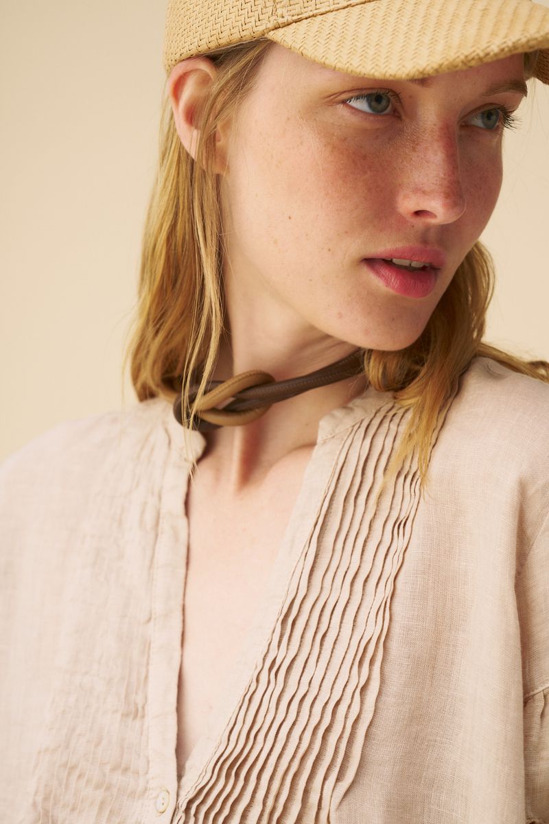 Close-up of a blond woman in a straw cap and tan linen blouse, wearing a knotted leather choker, with a neutral background.