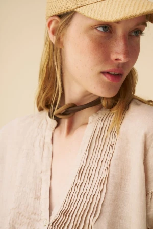 Close-up of a blond woman in a straw cap and tan linen blouse, wearing a knotted leather choker, with a neutral background.