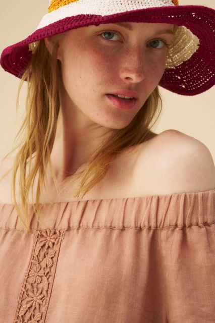 Close-up portrait of a fair-skinned young woman wearing a colorful straw hat and off-the-shoulder linen top, soft lighting, neutral background.