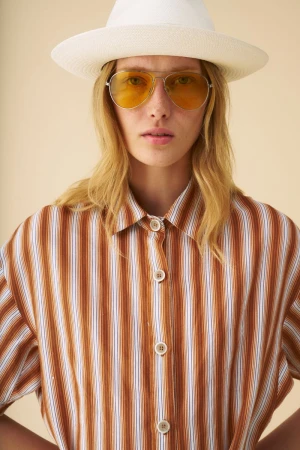 Model wearing a white fedora hat, yellow tinted aviator sunglasses, and a brown striped button-down shirt against a neutral background.