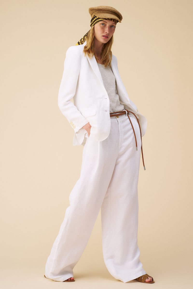 Fashion shot: Woman in a white linen suit and straw cap stands against a neutral background, exuding casual elegance and summery style.
