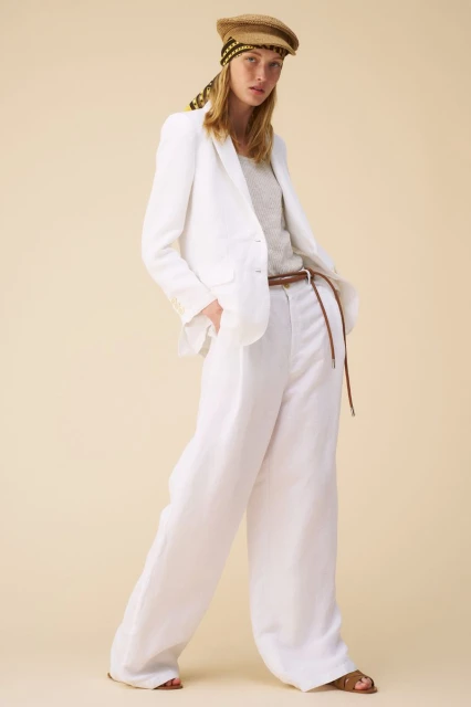 Fashion shot: Woman in a white linen suit and straw cap stands against a neutral background, exuding casual elegance and summery style.