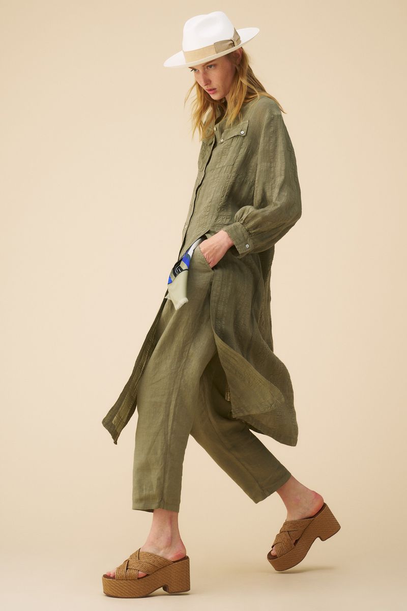An adult female model in a green linen outfit and white fedora hat walks in front of a neutral background, wearing brown platform sandals.
