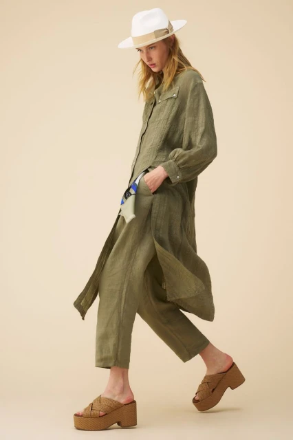 An adult female model in a green linen outfit and white fedora hat walks in front of a neutral background, wearing brown platform sandals.