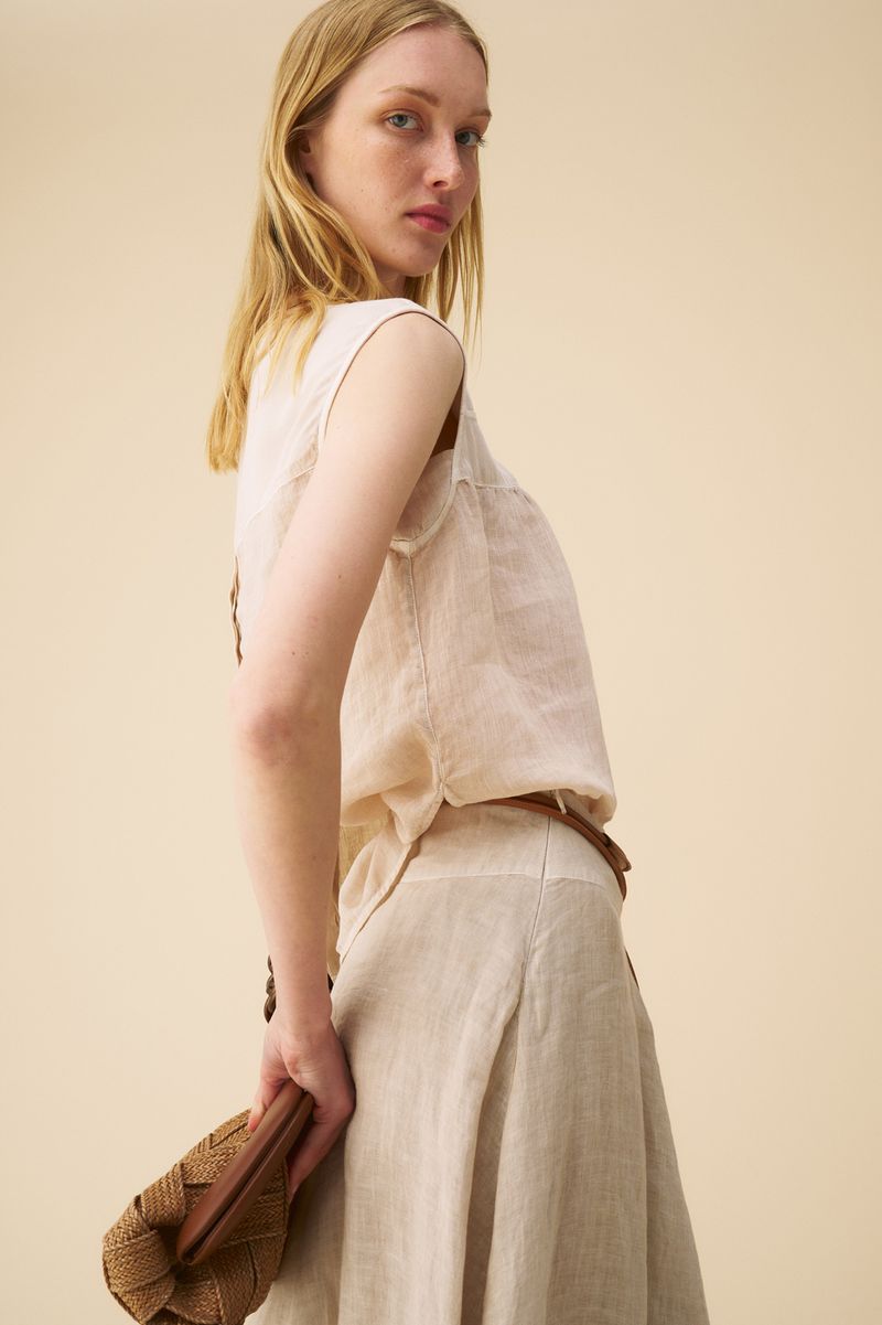 Woman in linen top and skirt holding a woven handbag, posing in a studio with a neutral backdrop, looking at the camera.