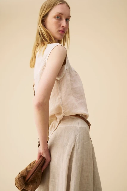 Woman in linen top and skirt holding a woven handbag, posing in a studio with a neutral backdrop, looking at the camera.