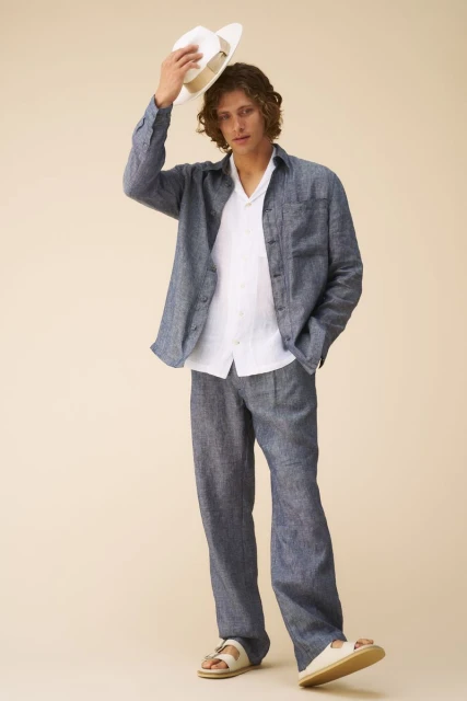 A slender adult male poses in a light-filled studio wearing a linen two-piece outfit, leather sandals, and a Panama hat.