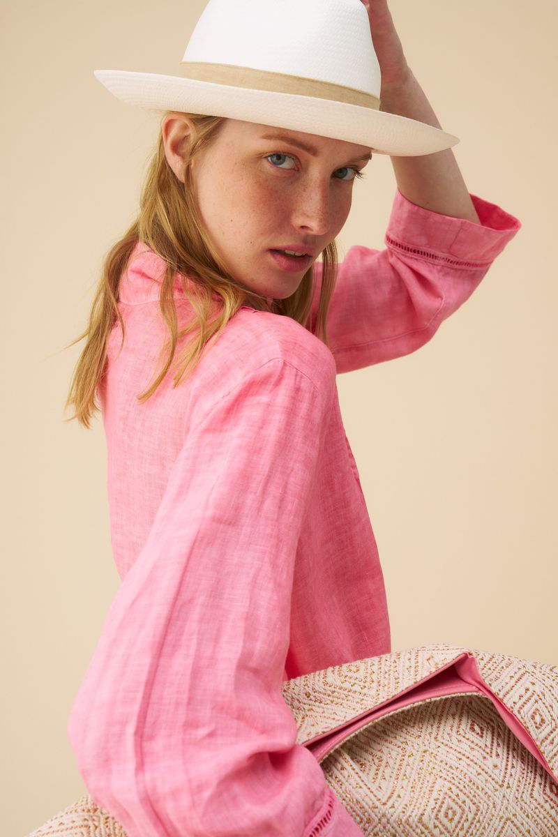 Portrait of a red-haired woman in a pink linen shirt and white straw hat, looking at the camera against a peach background.