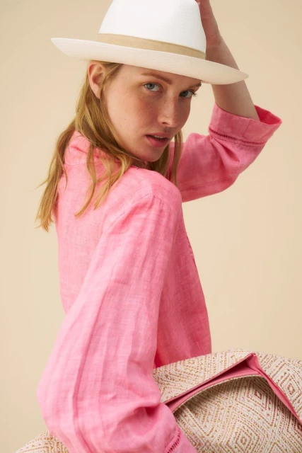 Portrait of a red-haired woman in a pink linen shirt and white straw hat, looking at the camera against a peach background.