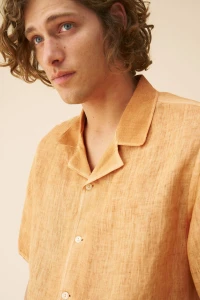Close-up portrait of a curly-haired man wearing an orange linen camp collar shirt against a neutral background.