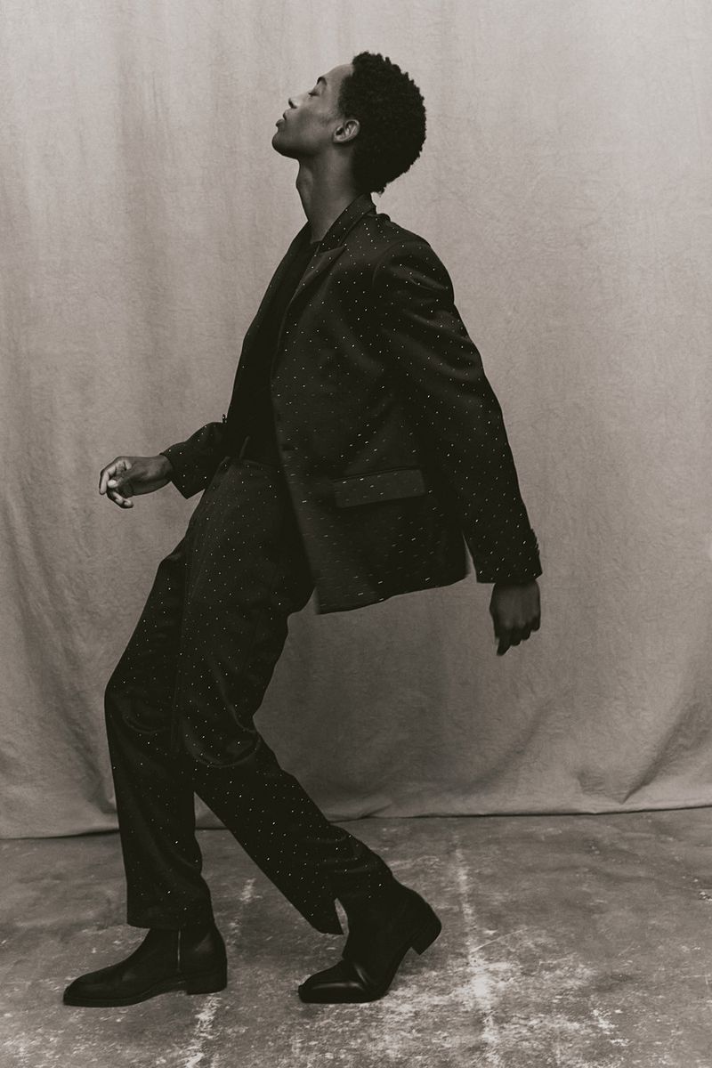 Monochrome studio shot of an African American male fashion model posing in a patterned suit and black boots against a neutral backdrop.