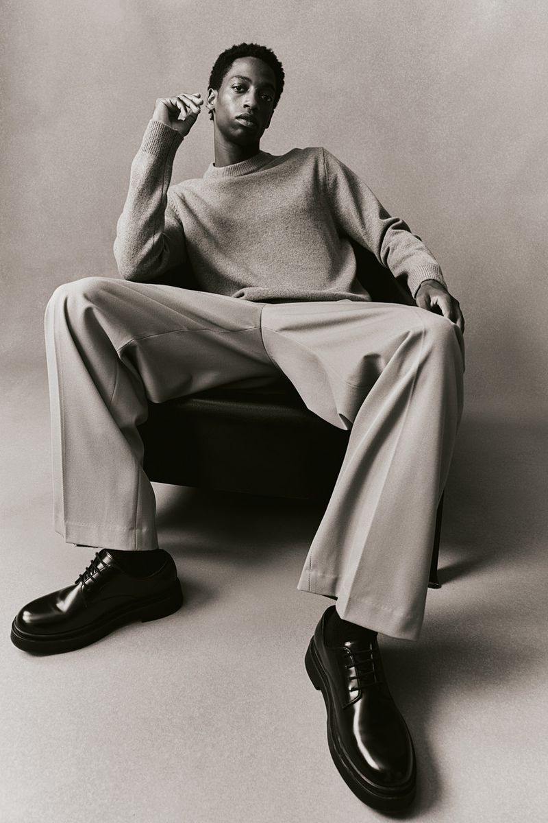 A sophisticated portrait of a Black male model in a grey sweater and khaki pants, seated in a chair against a grey backdrop.