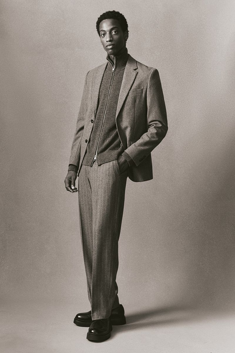 A young Black man is posed in a sepia-toned studio shot, wearing a grey suit, zip-up cardigan, and platform loafers, hand in his pocket.