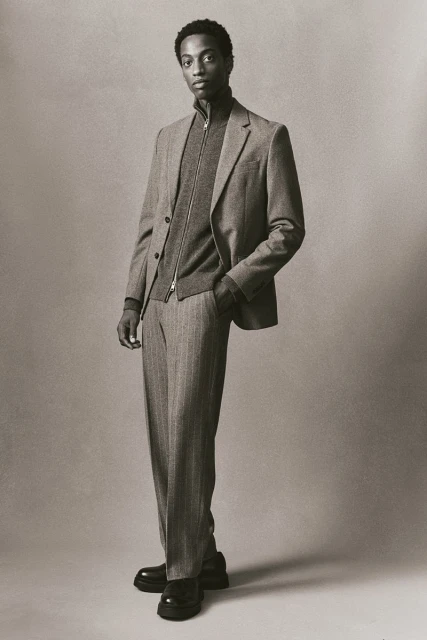 A young Black man is posed in a sepia-toned studio shot, wearing a grey suit, zip-up cardigan, and platform loafers, hand in his pocket.