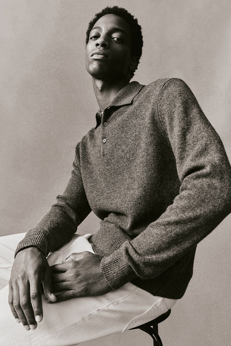 Portrait of a Black male model in a grey collared knit shirt and white pants, seated on a stool against a neutral studio backdrop.
