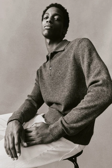 Portrait of a Black male model in a grey collared knit shirt and white pants, seated on a stool against a neutral studio backdrop.