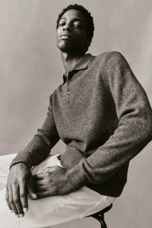 Portrait of a Black male model in a grey collared knit shirt and white pants, seated on a stool against a neutral studio backdrop.