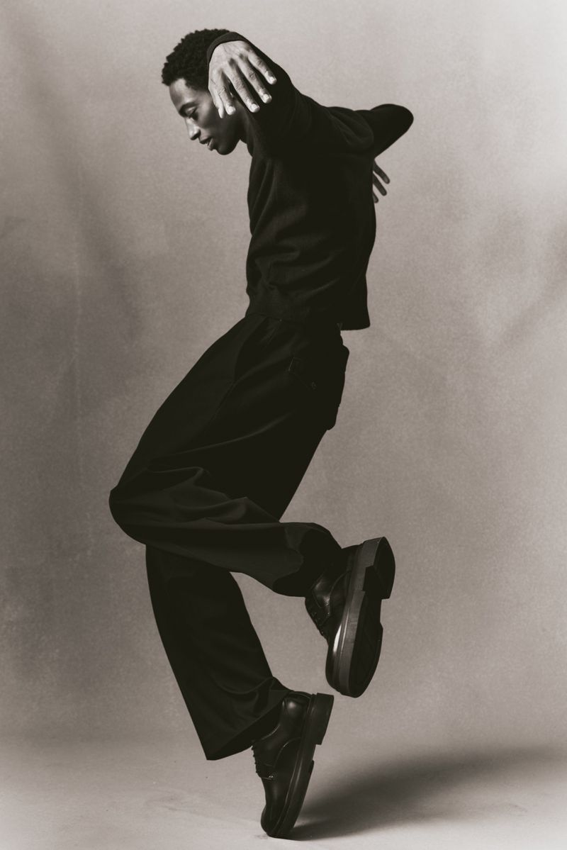 A Black male dancer in motion, wearing black clothing against a grey background, showcasing a dynamic pose and artistic expression.