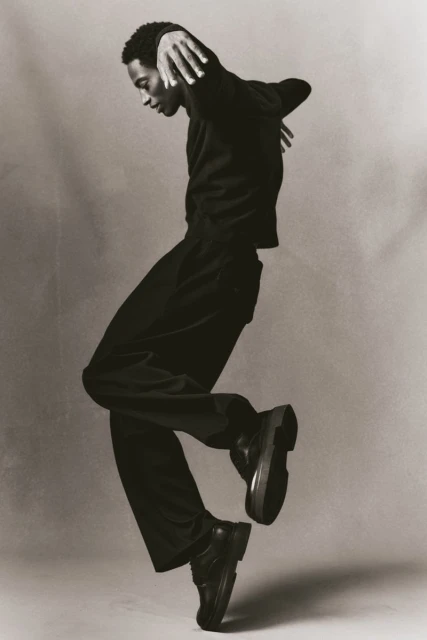 A Black male dancer in motion, wearing black clothing against a grey background, showcasing a dynamic pose and artistic expression.
