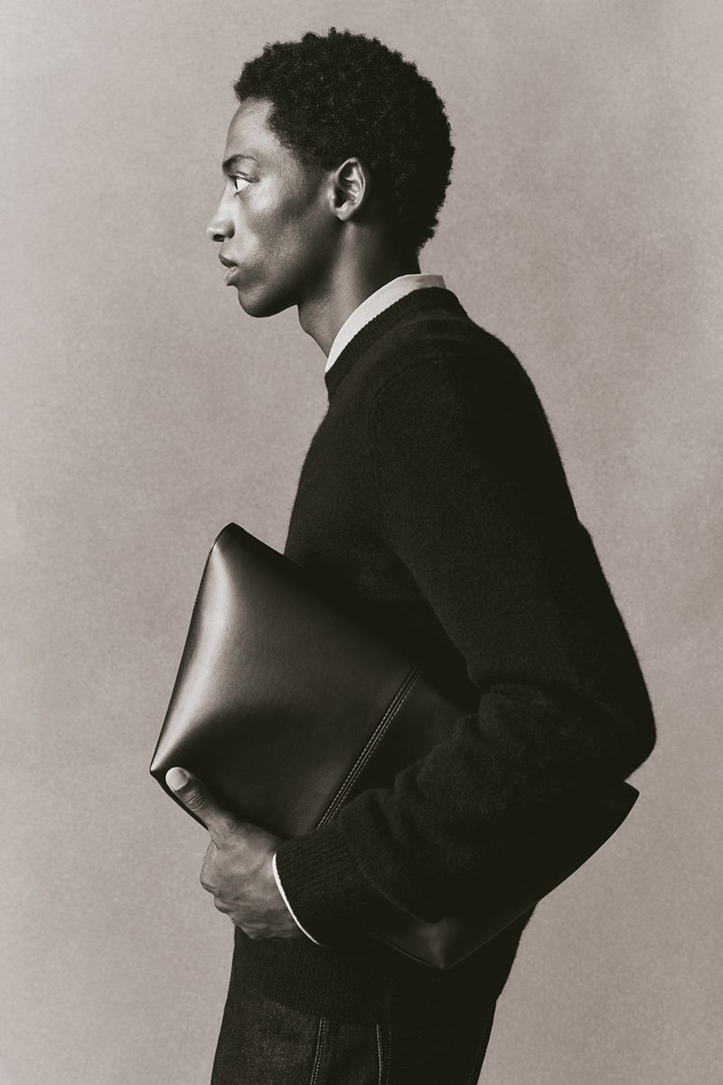 Profile shot of a Black male model in a sweater holding a leather clutch against a plain background in a studio setting with soft lighting.