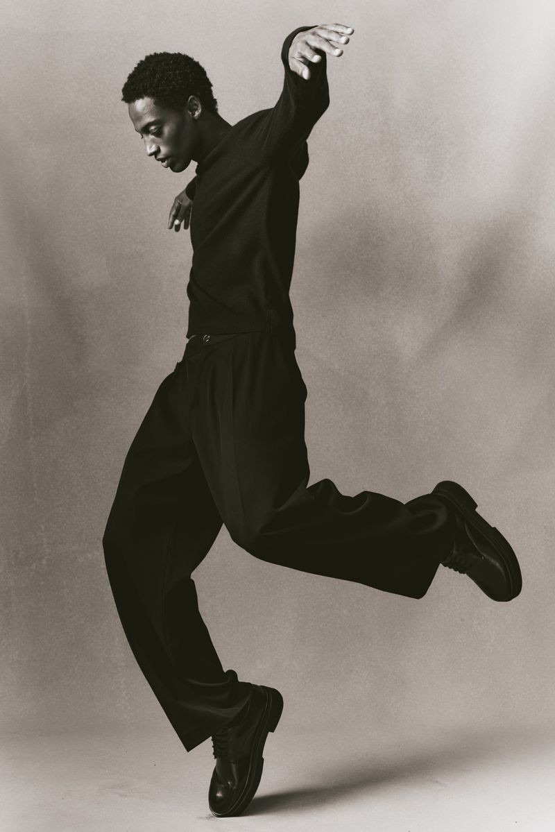 Black and white portrait of an African-American male dancer in black clothing, mid-air in a dynamic pose, against a neutral backdrop.