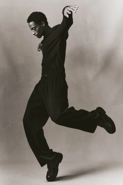 Black and white portrait of an African-American male dancer in black clothing, mid-air in a dynamic pose, against a neutral backdrop.