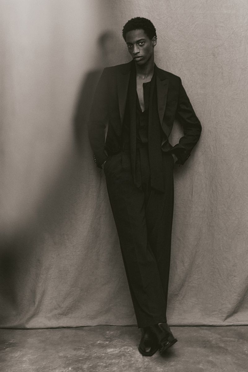 Model in a black suit leaning against a textured backdrop for an H and M project
