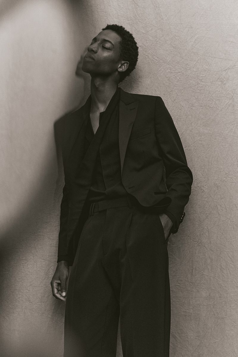 Model in dark suit leaning against textured wall with eyes closed in monochromatic fashion portrait