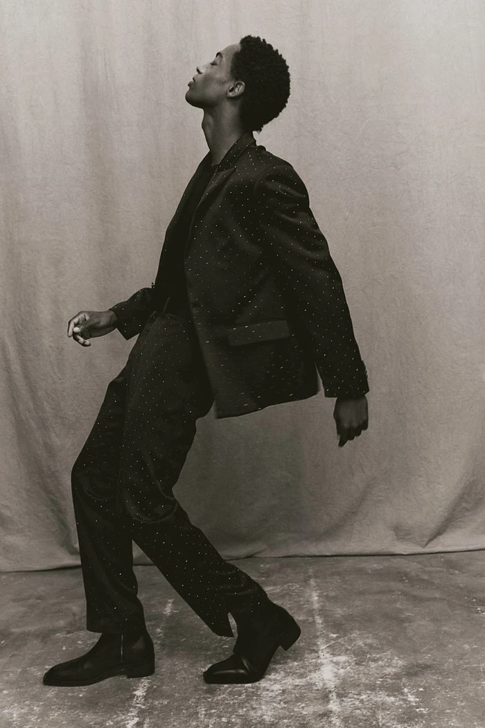 Model in a dark tailored suit posing dynamically against a neutral textured studio backdrop.