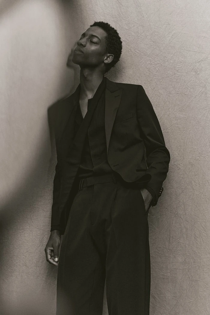 Model in dark suit leaning against textured wall with eyes closed in monochromatic fashion portrait