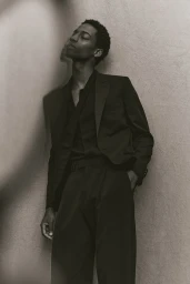 Model in dark suit leaning against textured wall with eyes closed in monochromatic fashion portrait