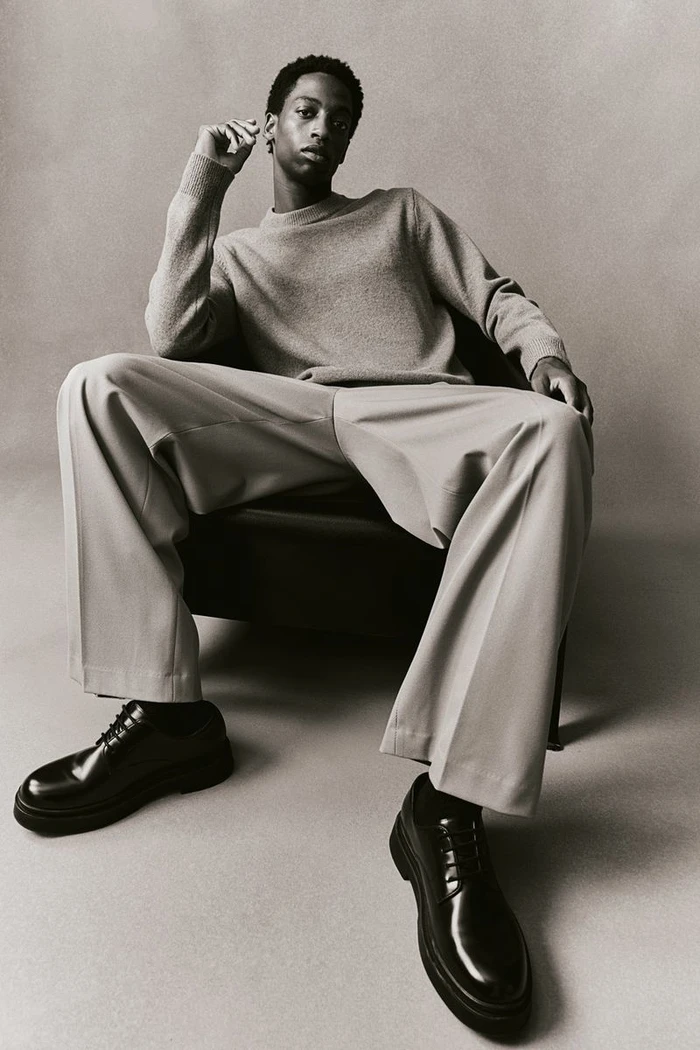 Black man seated in a dark chair wearing a grey sweater, light trousers, and black leather shoes against a grey background.