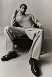 Black man seated in a dark chair wearing a grey sweater, light trousers, and black leather shoes against a grey background.