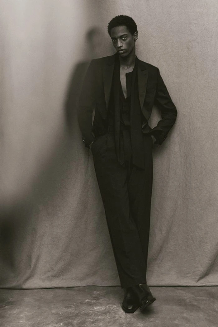 Model in a black suit leaning against a textured backdrop for an H and M project