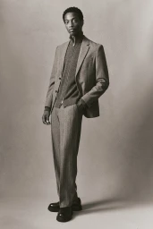 Model standing wearing a grey blazer, zip-neck sweater, and trousers against a neutral studio backdrop