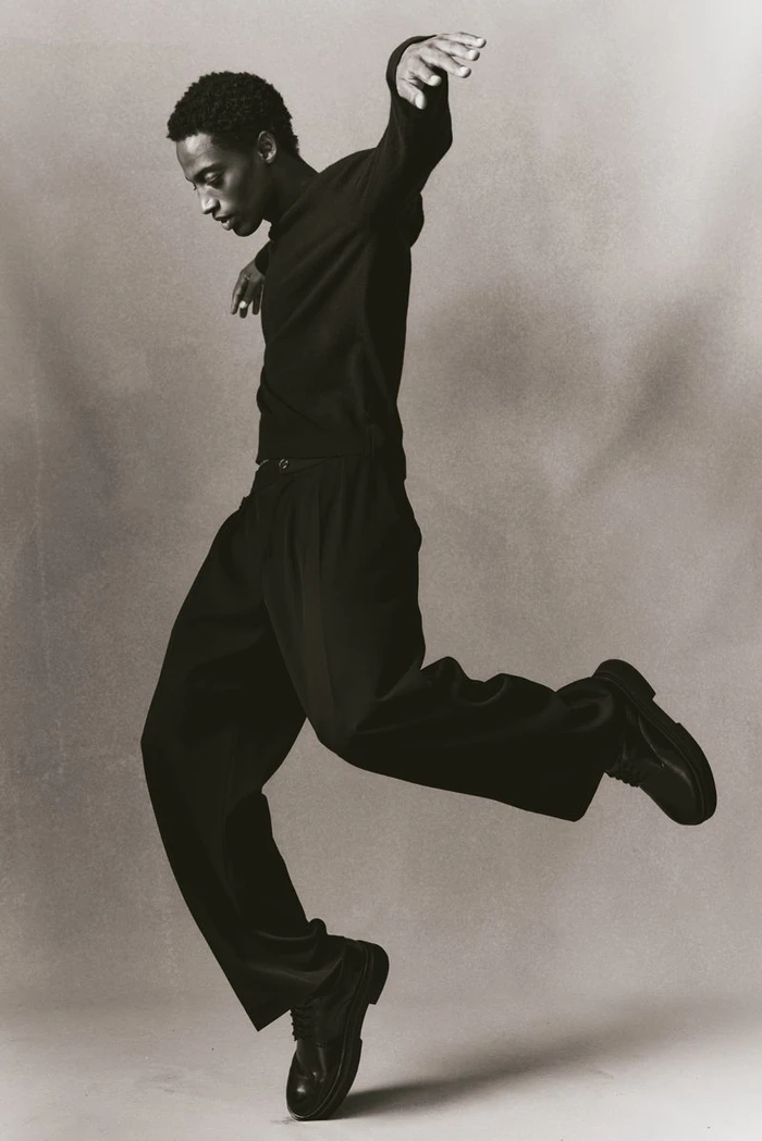 Black man in dark long-sleeve shirt and trousers leaping gracefully in studio