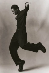 Black man in dark long-sleeve shirt and trousers leaping gracefully in studio
